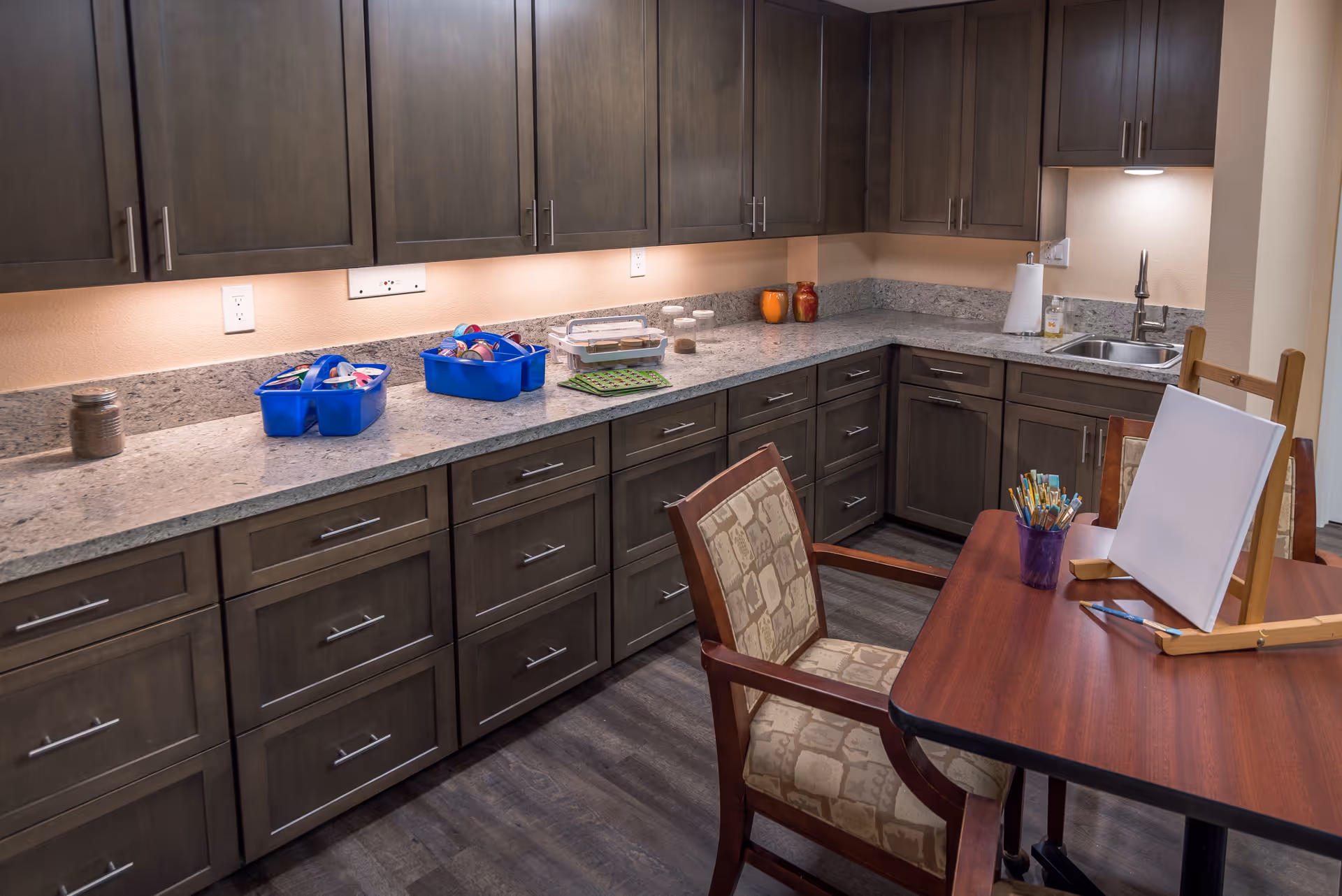Small kitchenette/activity room with a long granite countertop, gray cabinets, a sink, and a table with an easel and art supplies.