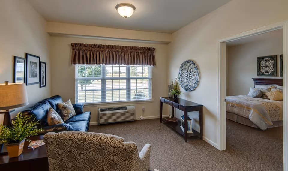 A bright living room with a sofa, armchair, console table and large window, with a bedroom visible through an open doorway.