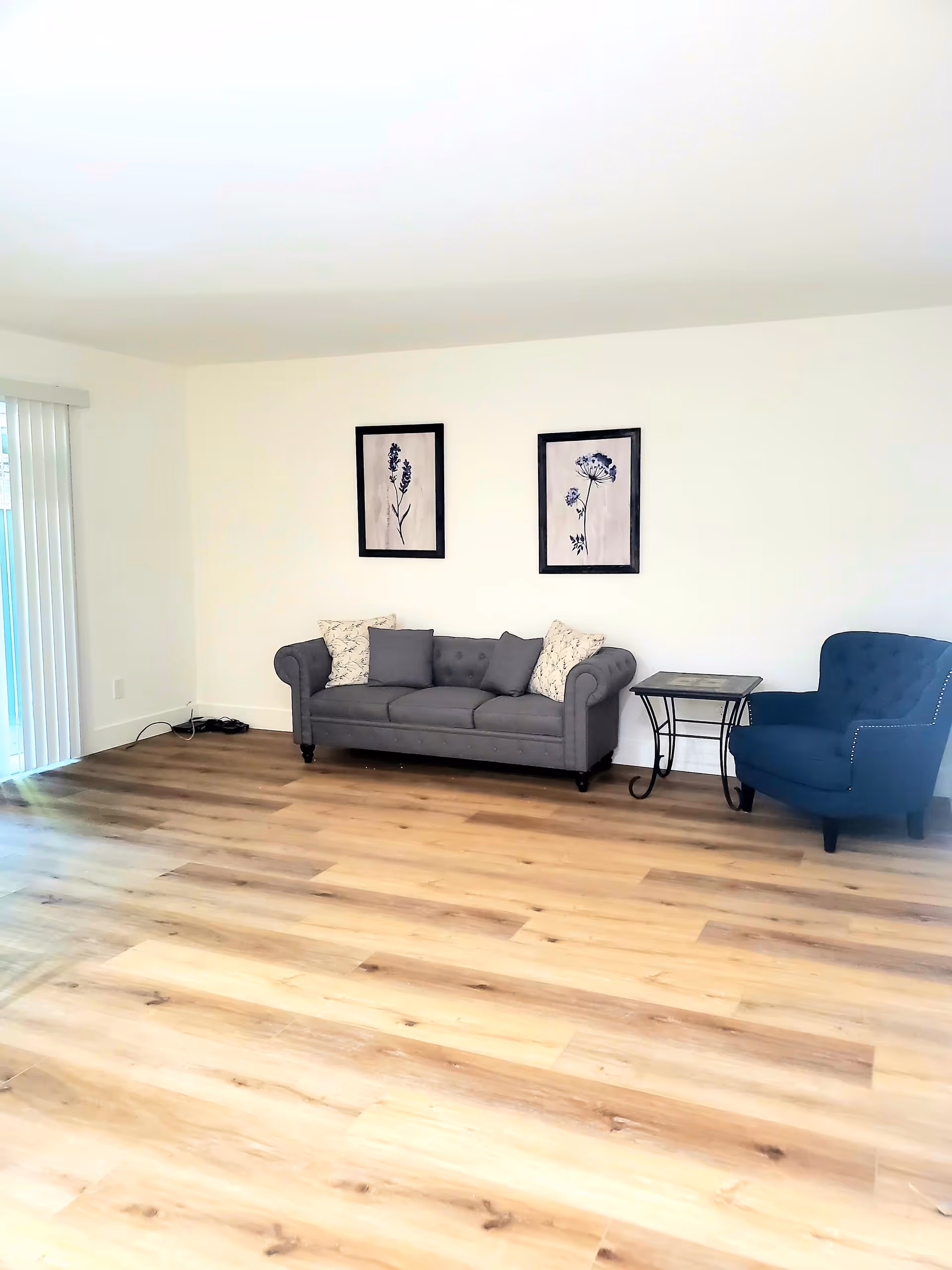 A living room with light wood flooring, a gray sofa with cushions, a blue armchair, a small side table, and two framed floral artworks on a white wall. There is a sliding glass door with vertical blinds on the left side.