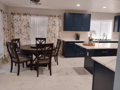 A modern kitchen and dining area featuring dark blue cabinets, white marble countertops, a round wooden dining table with six chairs, floral curtains on windows, and white tiled flooring.
