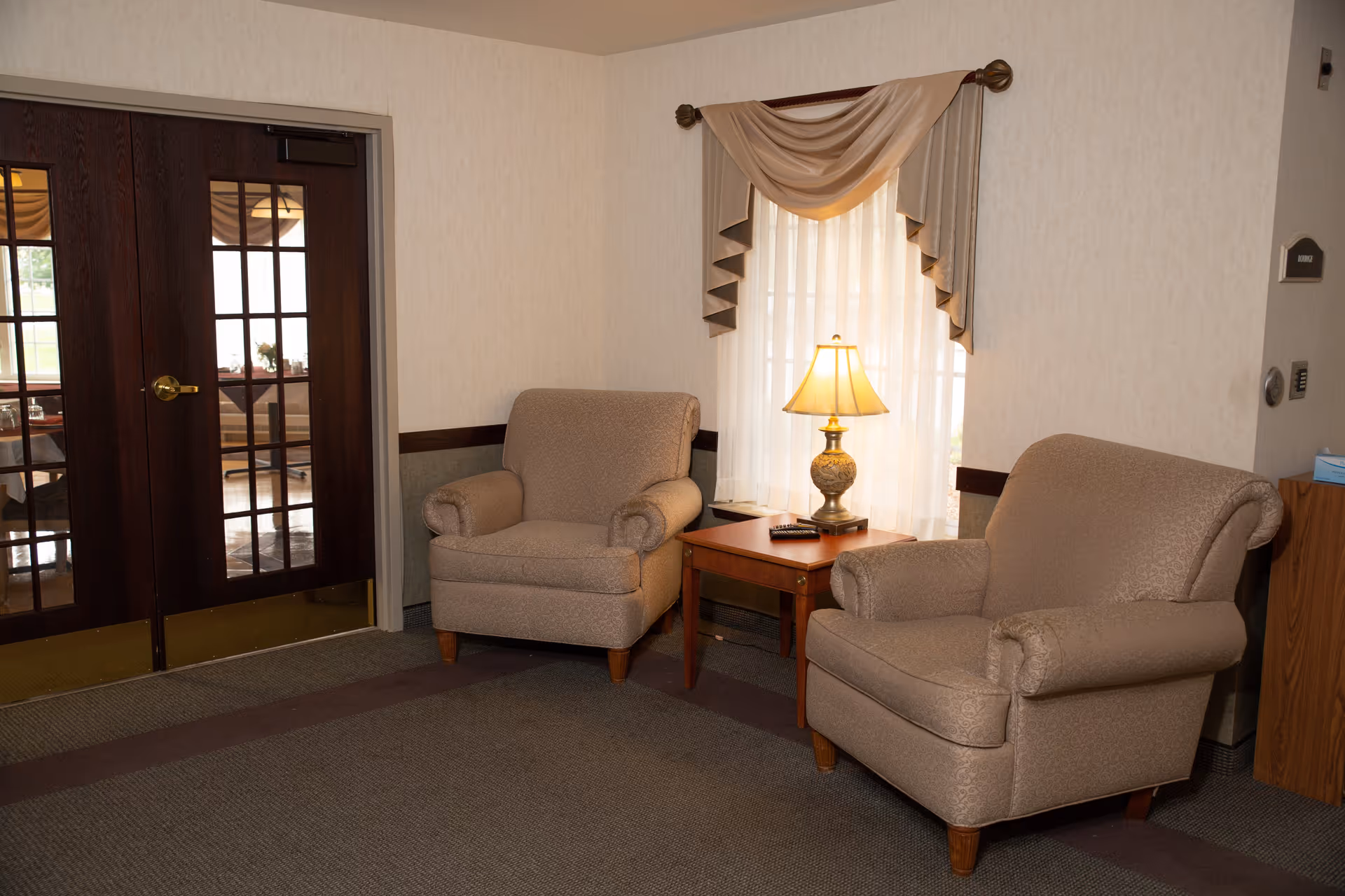 Two beige armchairs flanking a wooden side table with a lamp in a sitting area next to glass-paneled double doors.