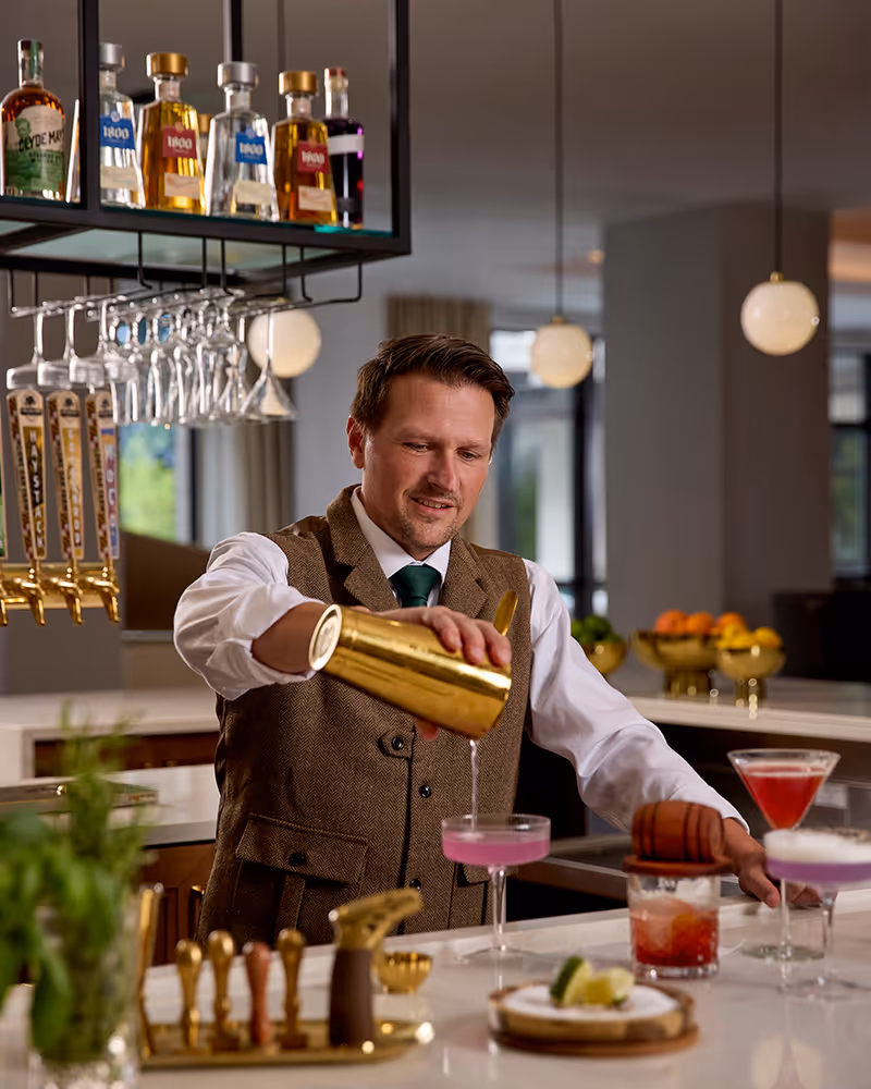 A bartender wearing a brown vest and white shirt is pouring a pink cocktail from a gold shaker into a coupe glass at a bar counter. The bar has various bottles and hanging glasses above, with several prepared cocktails and garnishes on the counter. The background shows a modern interior with pendant lights and large windows.