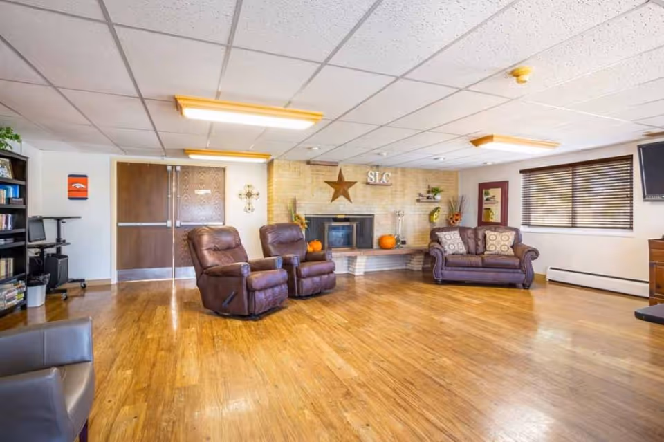 A spacious living room with wooden flooring, two brown recliner chairs, a brown couch with patterned cushions, a fireplace decorated with pumpkins and a star, a bookshelf with books and a computer station, and a window with blinds.