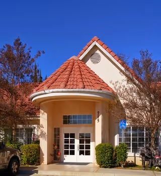Entrance of a building with a beige exterior and a red tiled roof under a clear blue sky. The entrance features double glass doors with white frames, flanked by windows and bushes on either side. The building number 715 is displayed near the door. There is a handicap parking sign visible to the right of the entrance.