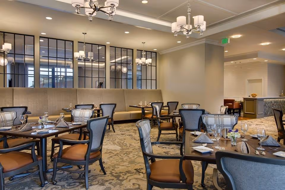 A well-lit dining room with multiple tables set with napkins, glasses, and silverware. The room features upholstered chairs with wooden frames, a long beige banquette along the wall, large mirrors with black grid frames, and elegant chandeliers hanging from the ceiling. The carpet has a patterned design, and the overall ambiance is warm and inviting.