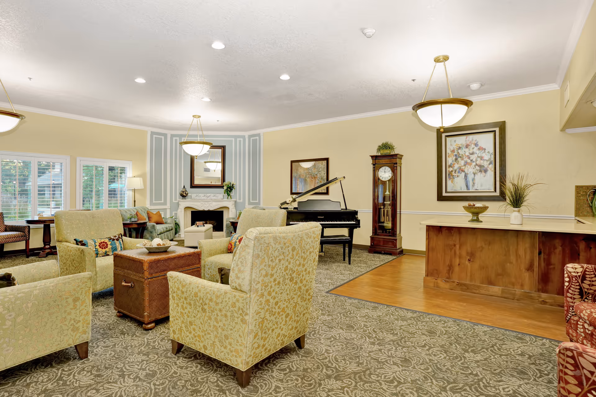 A spacious and well-lit living room area in a senior living facility with several upholstered armchairs arranged around a wooden chest used as a coffee table. The room features a grand piano, a grandfather clock, a fireplace with a mirror above it, and framed artwork on the walls. There are large windows with white shutters allowing natural light to enter, and the floor is covered with patterned carpet and a section of hardwood near a counter with decorative plants and a bowl.