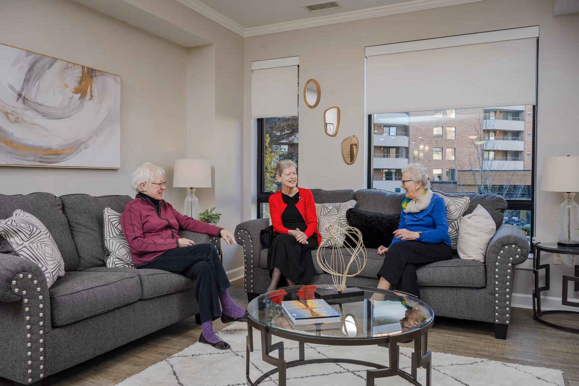 Three elderly women sitting and chatting on two gray sofas in a well-lit living room with large windows, a glass coffee table, decorative pillows, a modern abstract painting on the wall, and table lamps on side tables.