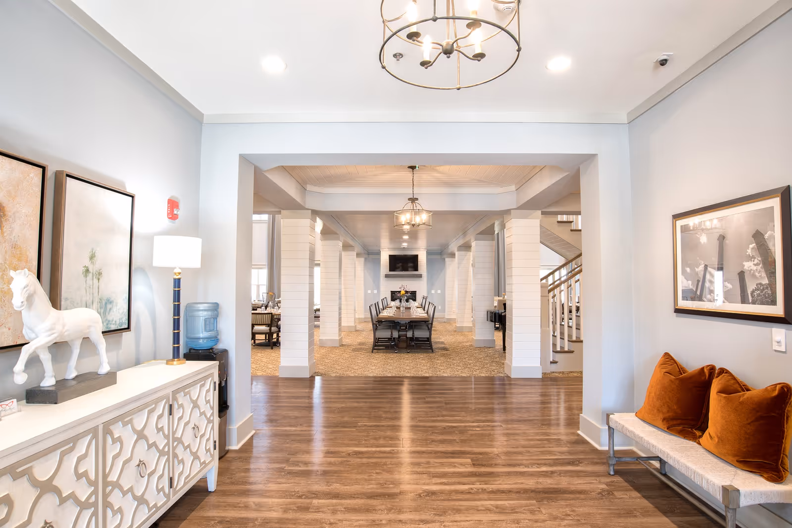 Interior view of a senior living facility showing a spacious hallway with wooden flooring leading to a dining area with a long table and chairs. The hallway features white pillars and ceiling lights. On the left side, there is a white decorative cabinet with a white horse statue and framed artwork above it. On the right side, there is a bench with two orange cushions and a framed black and white photograph above it.