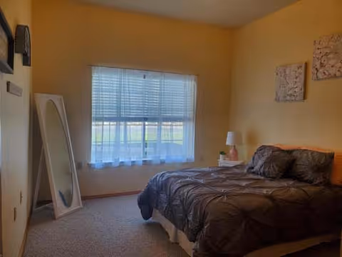 A bedroom with a large window covered by blinds and sheer curtains, a bed with a dark comforter and pillows, a white nightstand with a lamp and a small plant, a full-length mirror leaning against the wall, and two decorative wall hangings above the bed.