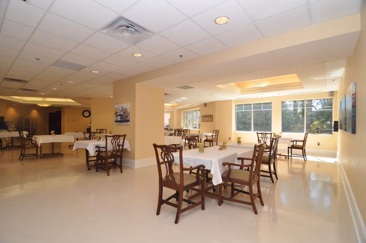 Bright dining room with multiple tables covered in white tablecloths and wooden chairs, lit by large windows and ceiling lights.