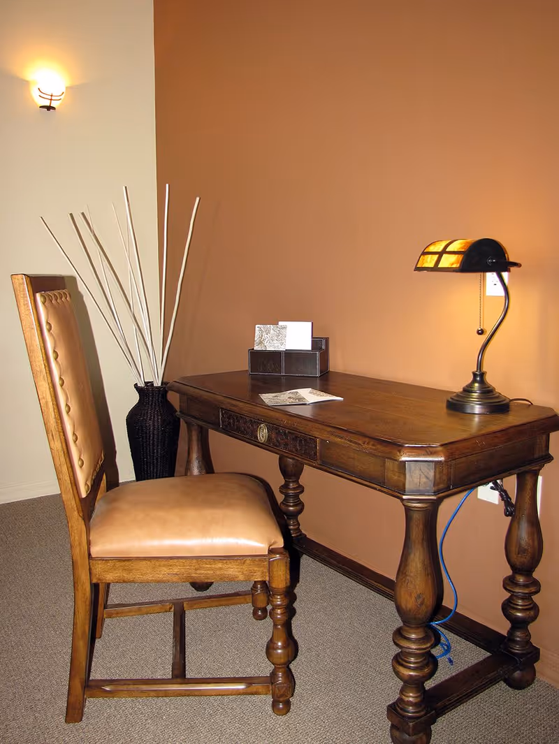 A wooden desk with ornate legs and a matching wooden chair with a padded leather seat and backrest. On the desk is a small decorative lamp with a pull chain, a card holder with cards, and a piece of paper. Behind the chair is a tall black vase with long white decorative sticks. The walls are painted in two tones: a light beige and a warm brown.