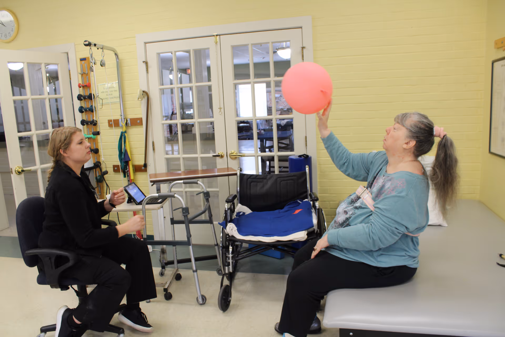 A woman sitting on a therapy table holding a pink balloon while a therapist seated on a chair watches her. There is a wheelchair, walker, and exercise equipment in the background in a room with yellow brick walls and glass-paneled double doors.