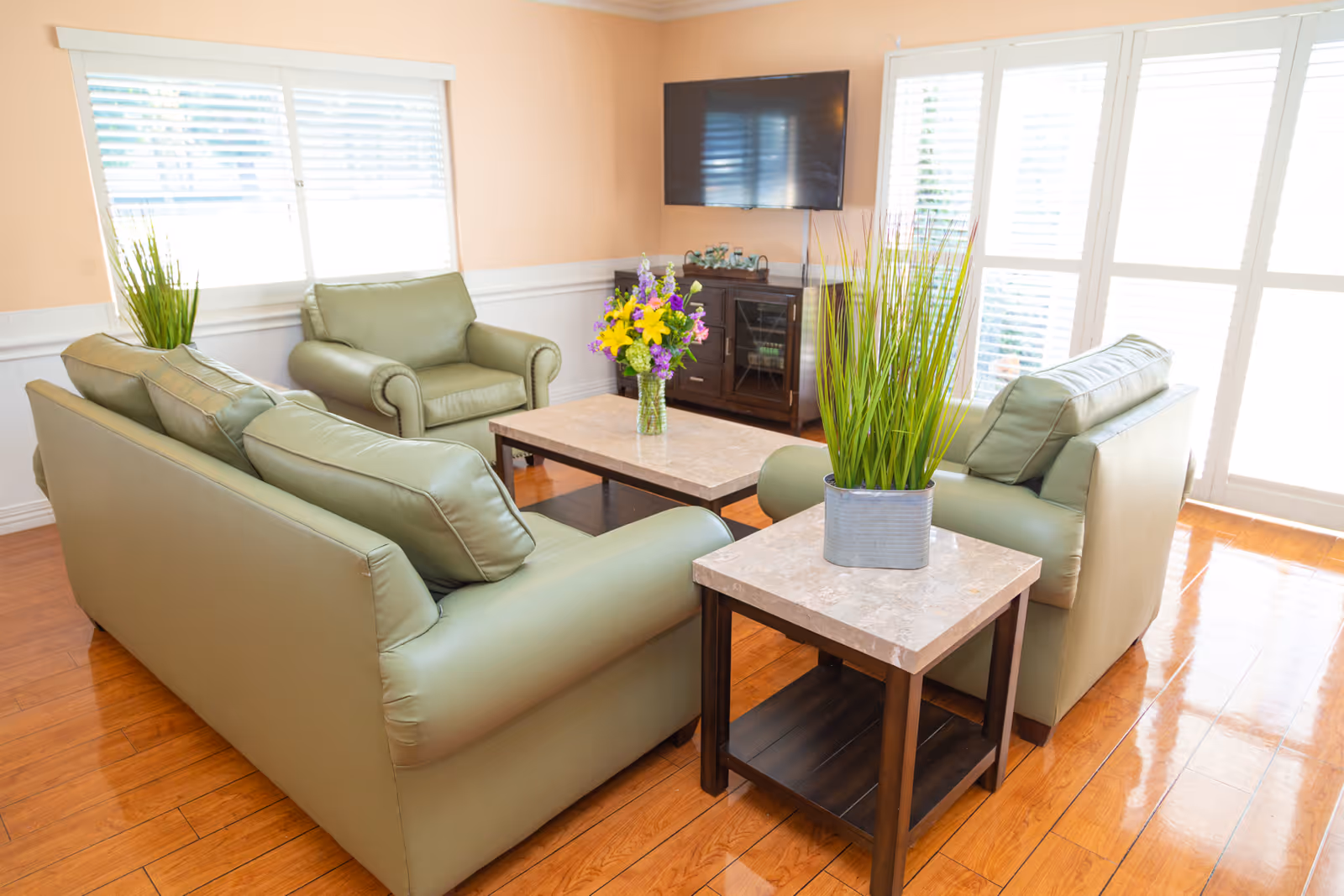 A bright living room with light green leather sofas and armchairs arranged around a marble-top coffee table and side table. The room has wooden flooring, large windows with white blinds, a flat-screen TV mounted on the wall, and decorative plants and flowers on the tables.
