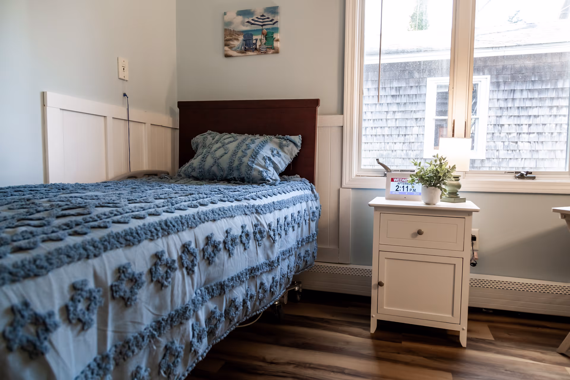 A single bed with a blue textured bedspread and matching pillow in a room with light blue walls and wood flooring. Next to the bed is a white nightstand with a small lamp, a potted plant, and a digital clock showing the time 2:11 PM. A window with a view of a shingled exterior wall is behind the nightstand. A small beach-themed painting hangs on the wall above the bed.