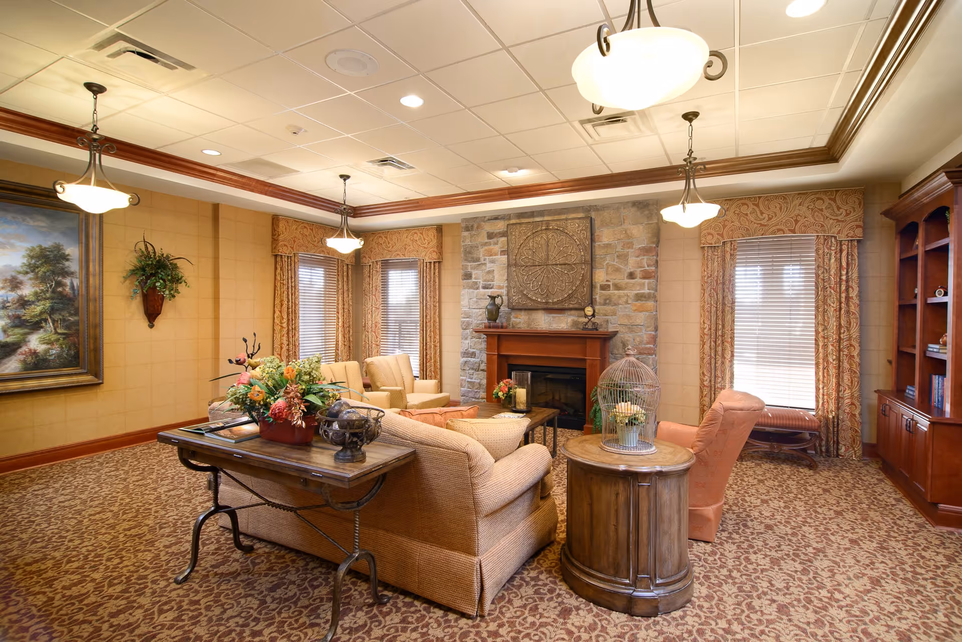 A cozy living room with patterned carpet and beige walls, featuring a stone fireplace with a decorative piece above it. The room has several upholstered chairs and sofas arranged around a wooden coffee table and side table with floral and decorative items. There are three hanging ceiling lights, large windows with patterned curtains, a wooden bookshelf, and a framed landscape painting on the wall.