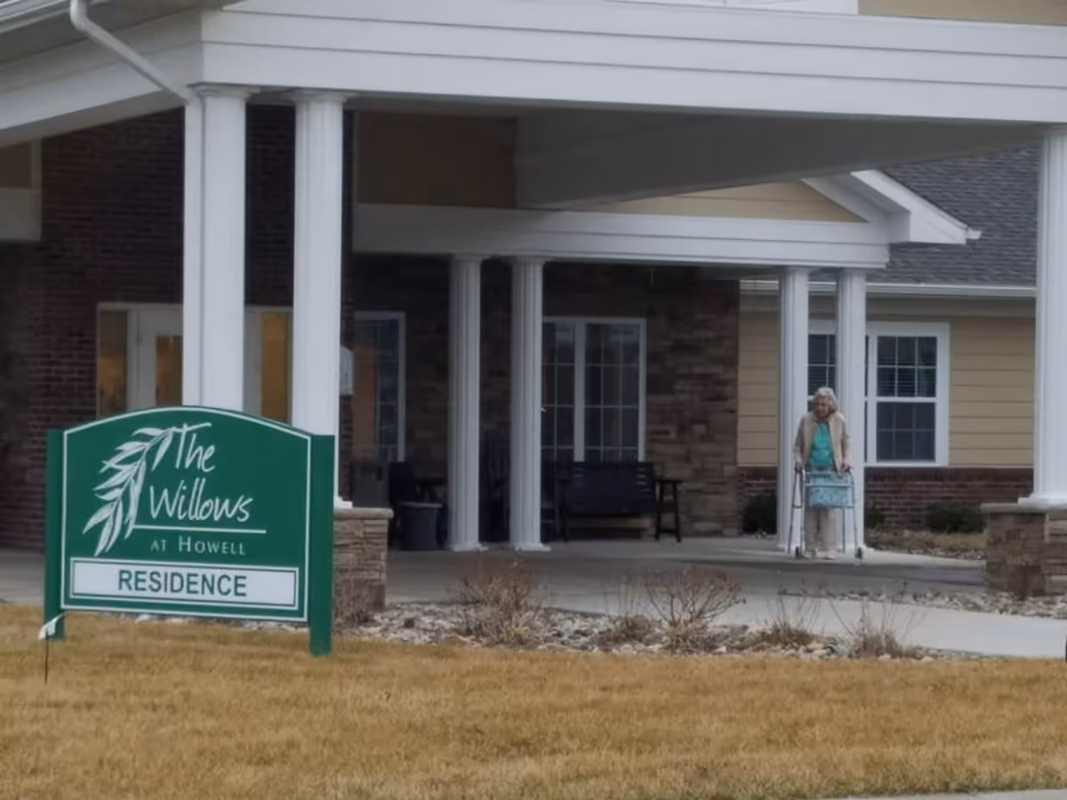 Entrance of The Willows At Howell residence with white pillars and a covered porch. An elderly woman using a walker is seen near the entrance. A green sign with white text reads 'The Willows At Howell RESIDENCE'.