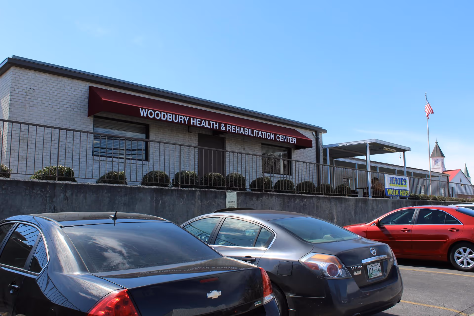 Front exterior of Woodbury Health & Rehabilitation Center with a red awning sign, parked cars in the foreground and an American flag.