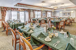 A spacious dining room with multiple tables covered in green tablecloths, each set with white napkins, cups, glasses, and silverware. The room has large windows with floral curtains allowing natural light to enter. The walls are decorated with framed pictures and the ceiling has round light fixtures.