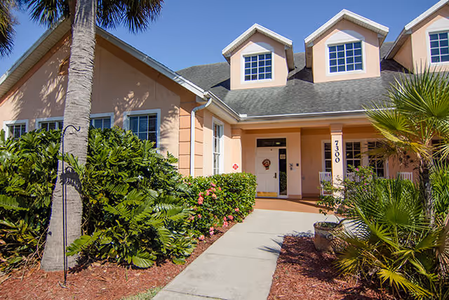 Exterior view of a peach-colored building with a gray roof, featuring three dormer windows. A concrete walkway leads to the entrance, which has a white door and a porch area with two rocking chairs. The building is surrounded by green shrubs, palm trees, and other landscaping under a clear blue sky.