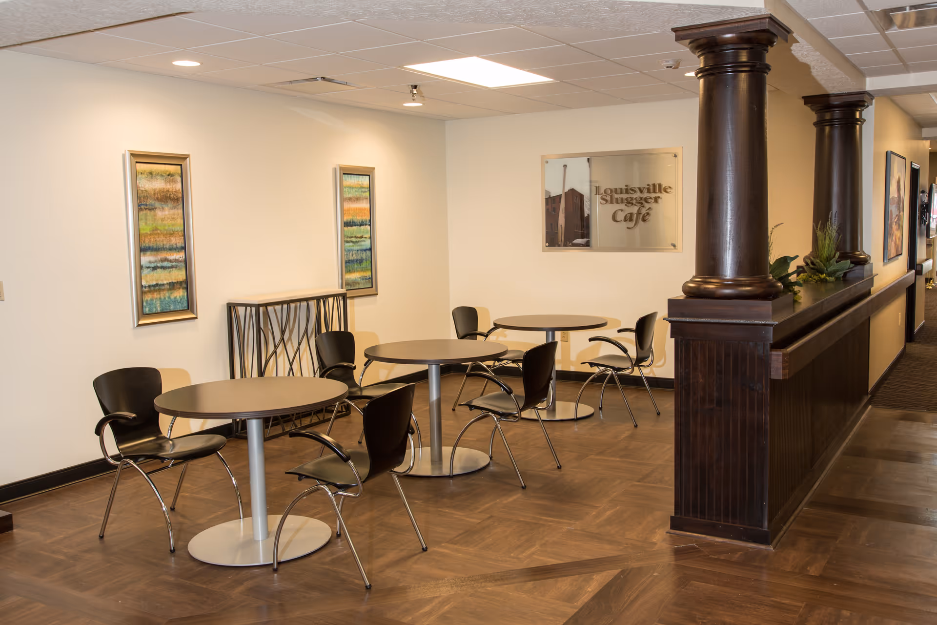 Interior view of a small café area with round tables and black chairs. The space features wooden flooring, two framed abstract paintings on the wall, and a sign that reads 'Louisville Slugger Café'. There are dark wooden columns and a half wall separating the café area from a hallway.