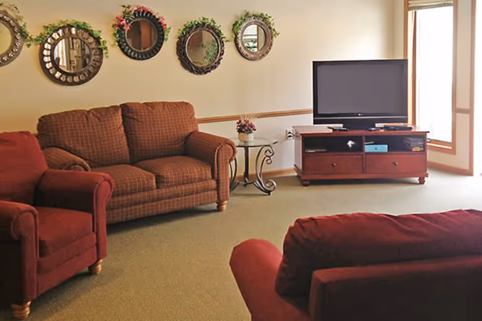 A cozy living room with a red armchair and a brown checkered loveseat. There is a wooden TV stand with a flat-screen television on it, and a small glass side table with a flower arrangement. Five decorative round mirrors with different frames are hung on the wall above the loveseat. The room has beige walls and carpeted floor, with large windows allowing natural light to enter.