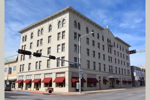 A large, multi-story beige building with red awnings over the ground floor windows, located at a street intersection with traffic lights and street signs visible under a clear blue sky.