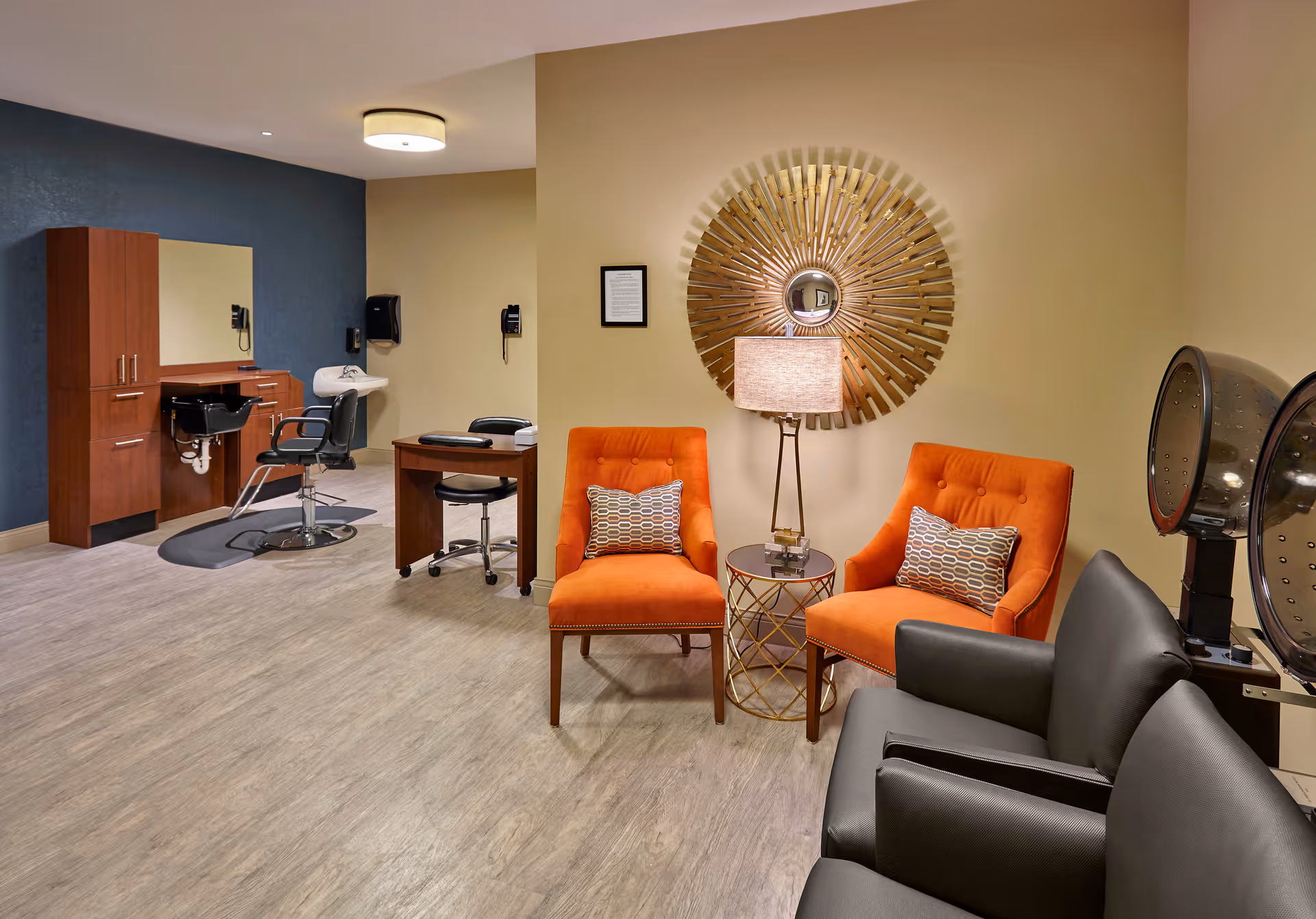 Interior view of a salon area in a senior living facility featuring two orange armchairs with patterned pillows, a small round table with a lamp, a decorative sunburst mirror on the wall, two black salon chairs, a wooden cabinet, a sink, and hair drying stations.