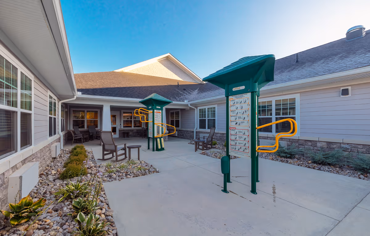 Outdoor courtyard area of The Springs at Wyandot Trail featuring exercise equipment with instructional signs, several chairs and small tables, surrounded by a building with multiple windows and a stone and siding exterior under a clear blue sky.