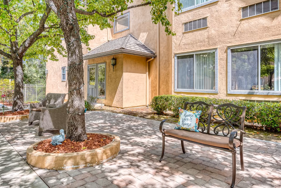 Outdoor patio area at Chino Hills Senior Living with a metal bench featuring a decorative backrest and a cushion with the words 'home sweet home'. There are two wicker chairs near a tree with string lights wrapped around its trunk. The patio is paved with stone tiles and surrounded by bushes and a beige building with windows and a door.
