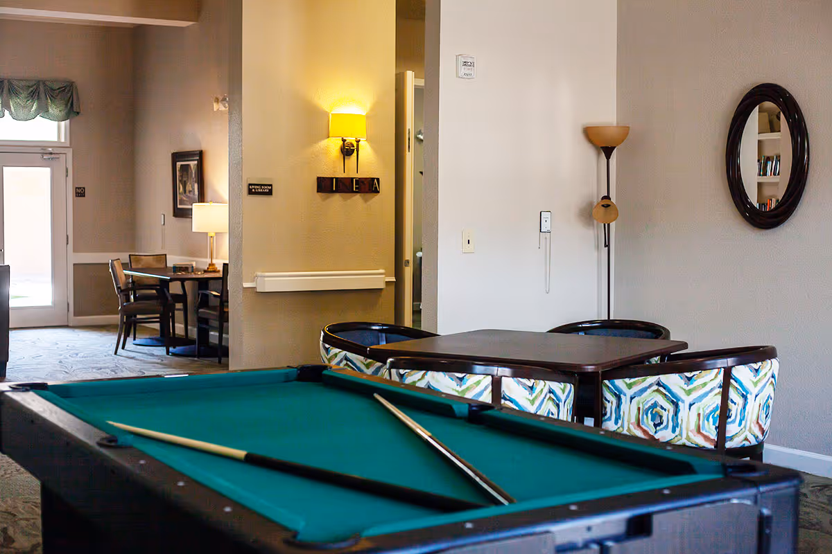 Interior view of a senior living facility common area featuring a pool table with two cues resting on it, a square table surrounded by four colorful patterned chairs, a floor lamp, a round wall mirror, and a glimpse of another seating area with chairs and a table near a door with a window.