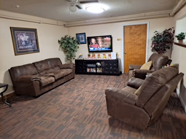 A cozy living room with brown leather sofas and recliners arranged around a black TV stand with a flat-screen television mounted on the wall. The room has beige walls, a patterned carpet, a wooden door, framed artwork, and two potted plants in the corners.