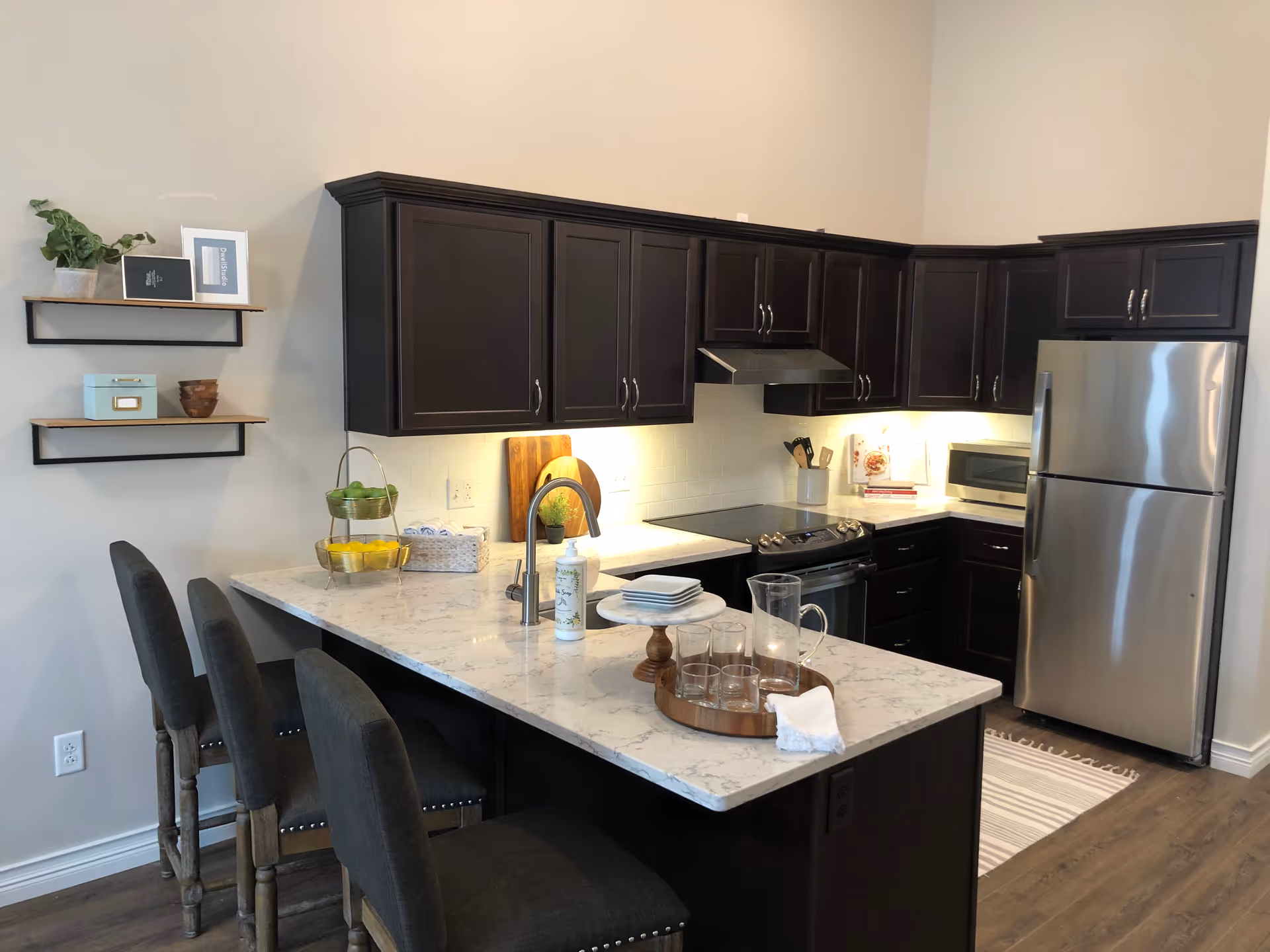 Modern kitchen with dark wood cabinets, stainless steel refrigerator and stove, white marble countertop island with three dark upholstered chairs, and decorative items including a tiered fruit basket, plates, glasses, and a pitcher.
