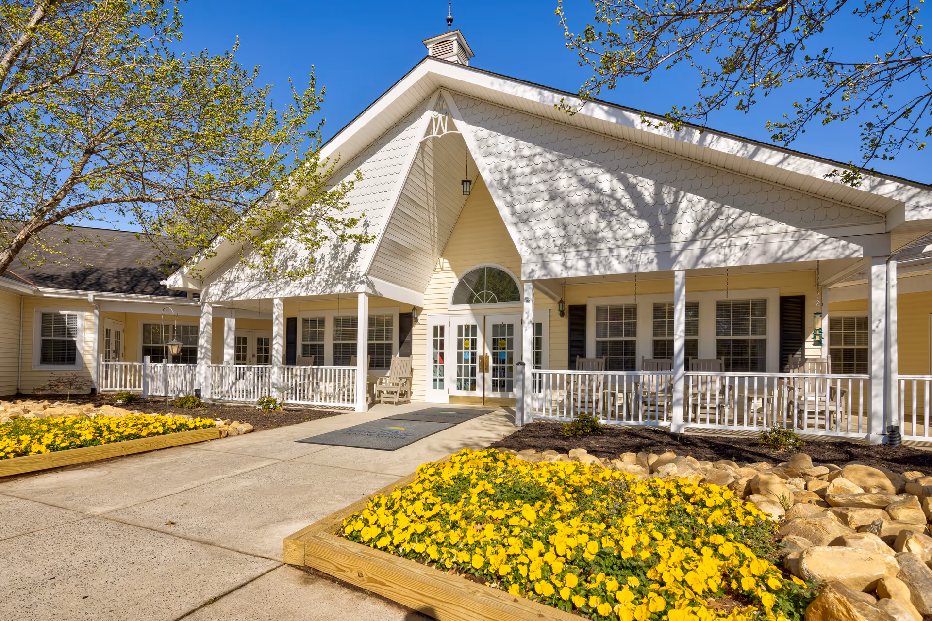 Front exterior view of a single-story building with a peaked roof and a covered porch. The entrance features double glass doors with sidelights and a semicircular window above. There are rocking chairs on the porch and flower beds with yellow flowers and rocks along the walkway leading to the entrance. Trees with budding leaves frame the scene under a clear blue sky.