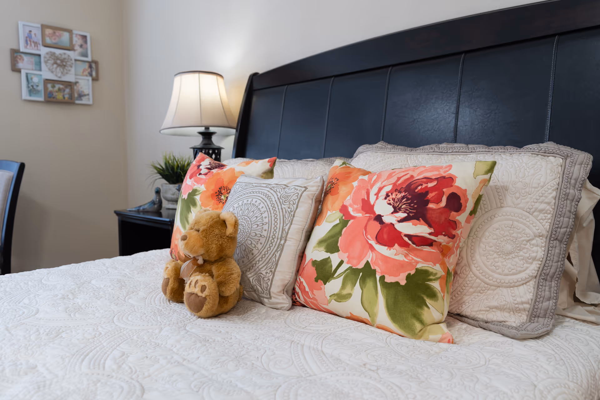 A neatly made bed with a black leather headboard, decorated with floral and patterned pillows, and a small teddy bear sitting on the bedspread. A bedside table with a lamp and a small plant is visible in the background, along with a wall decorated with framed pictures.