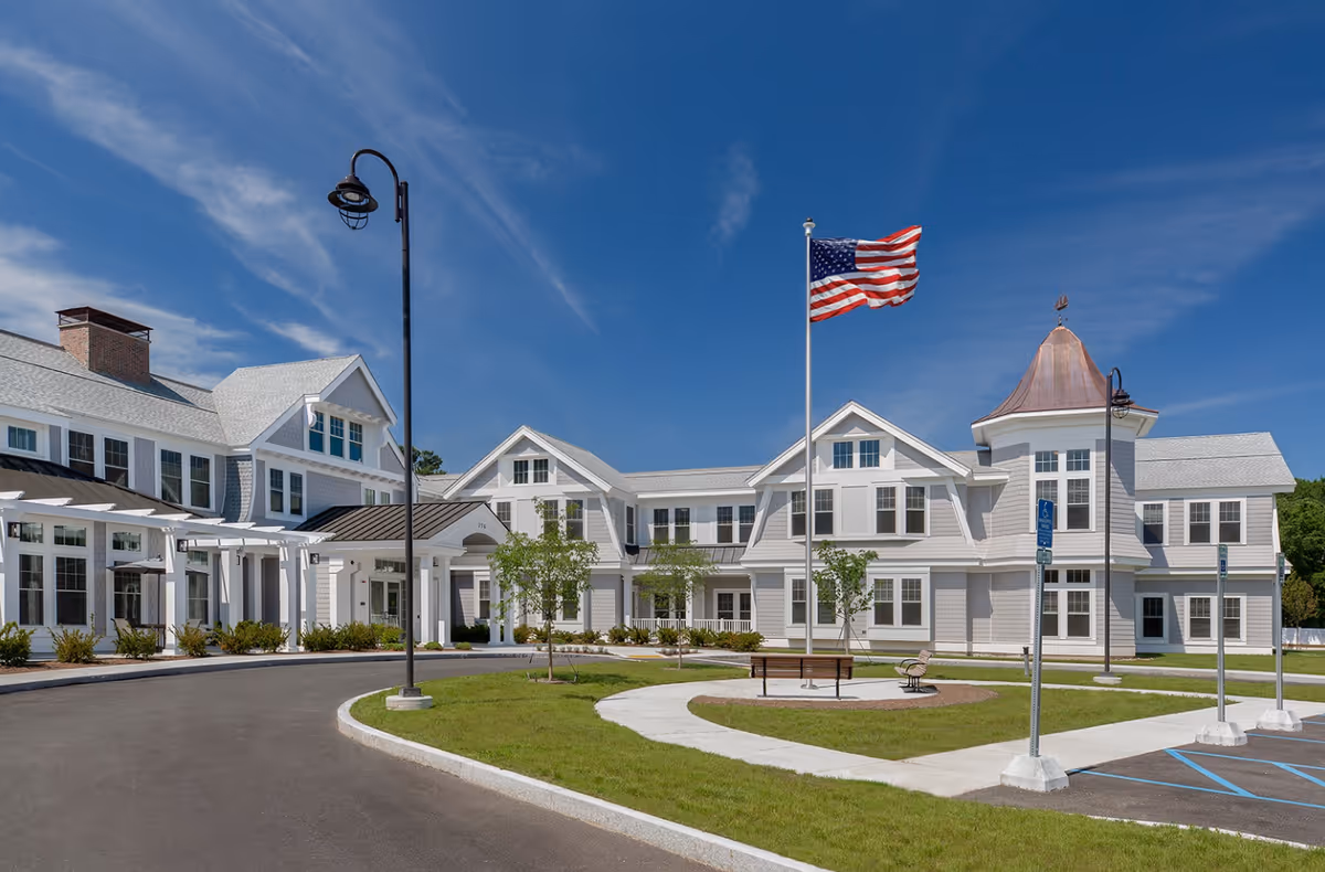 Exterior view of a large, white senior living facility building with multiple windows, a covered entrance, a curved driveway, a flagpole with an American flag, benches, and a clear blue sky.