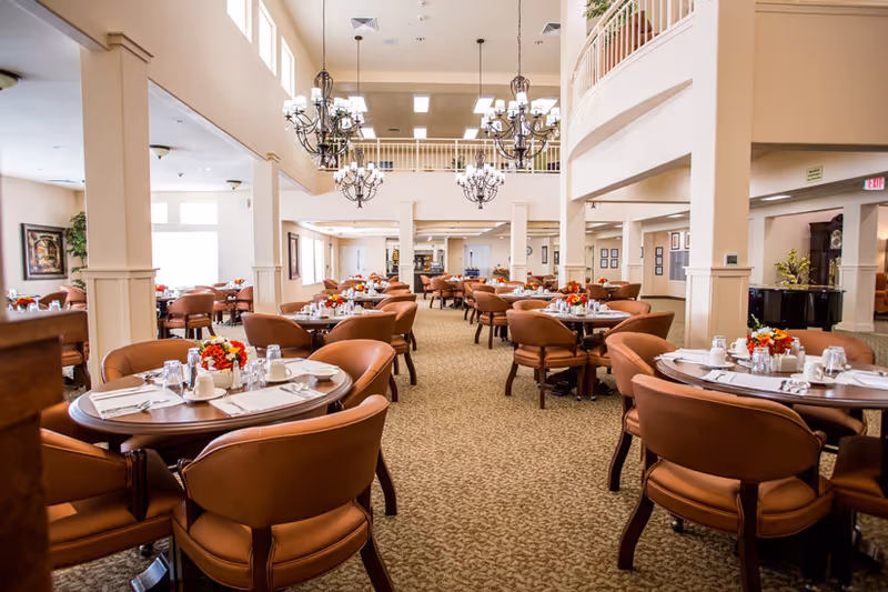 Spacious two-story dining room set with round tables, upholstered chairs, chandeliers, and floral centerpieces.