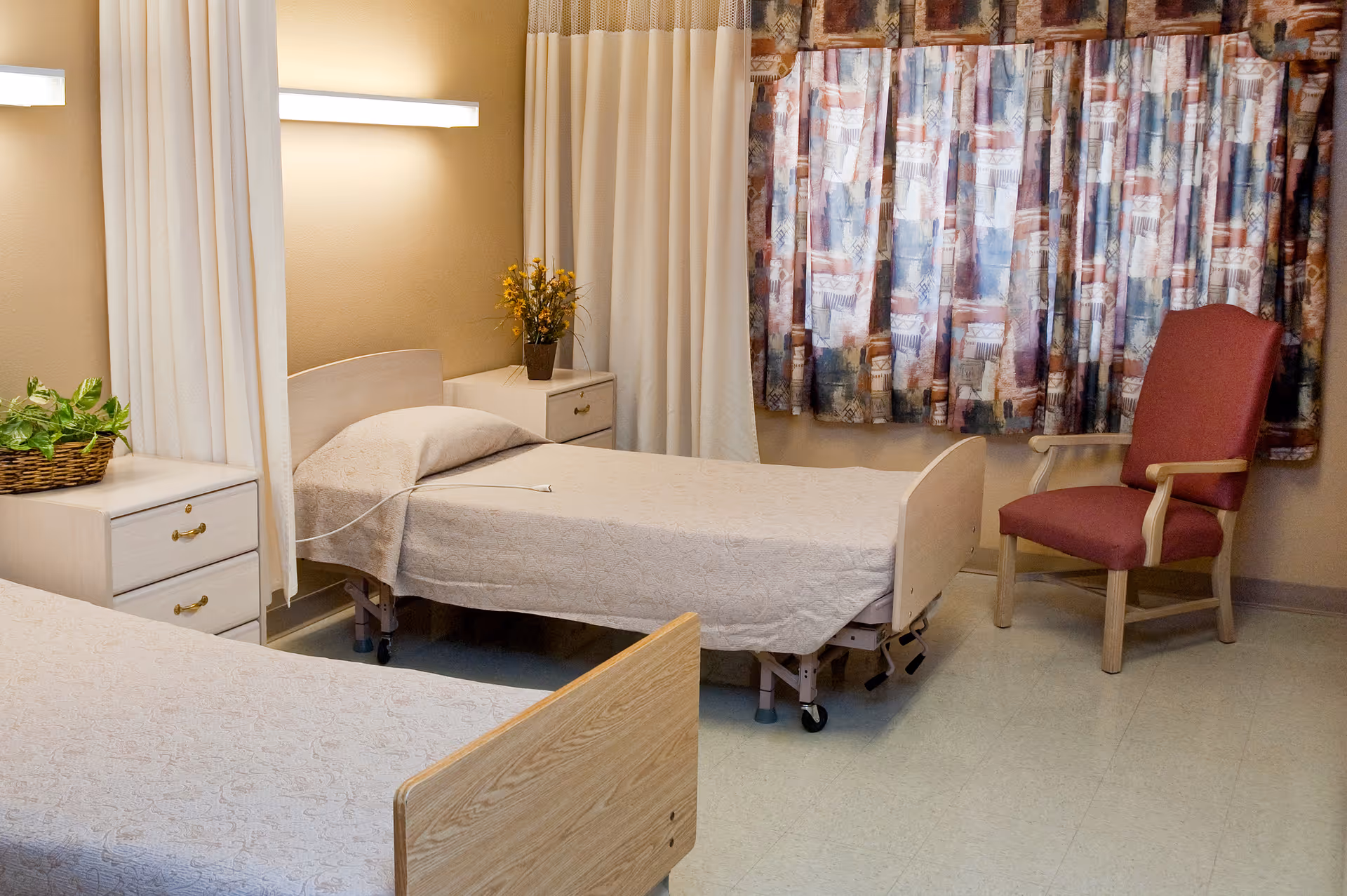 A room with two hospital-style beds covered with beige bedspreads, a bedside table with a plant and a vase with yellow flowers, a red armchair, and patterned curtains covering a window.