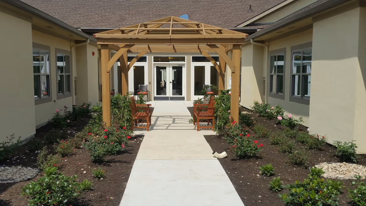 Outdoor courtyard area with a wooden pergola over a concrete pathway, surrounded by flower beds with blooming roses and green shrubs. There are wooden benches under the pergola and a building with multiple windows on both sides.