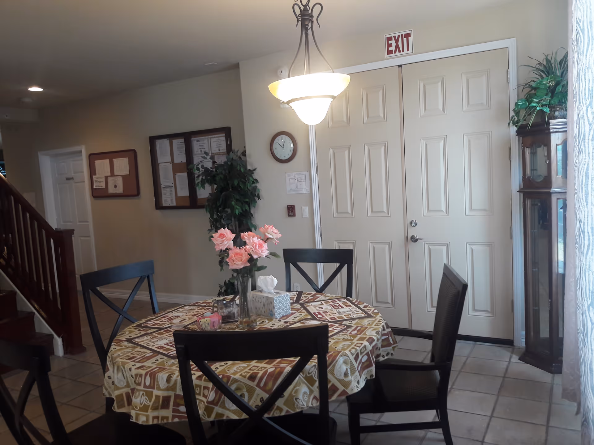 Interior view of a common area with a round table covered with a patterned tablecloth and four chairs around it. A vase with pink flowers and a tissue box are on the table. There is a large double door with an exit sign above it, a clock on the wall, a bulletin board, a potted plant, and a wooden grandfather clock near a window with sheer curtains.