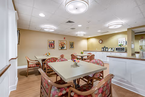 A well-lit dining area with several tables and cushioned chairs featuring a red and beige patterned upholstery. The room has light-colored walls adorned with framed floral artwork. In the background, there is a counter with coffee and beverage dispensers, a microwave, and a clock on the wall. The ceiling has recessed lighting and circular light fixtures.