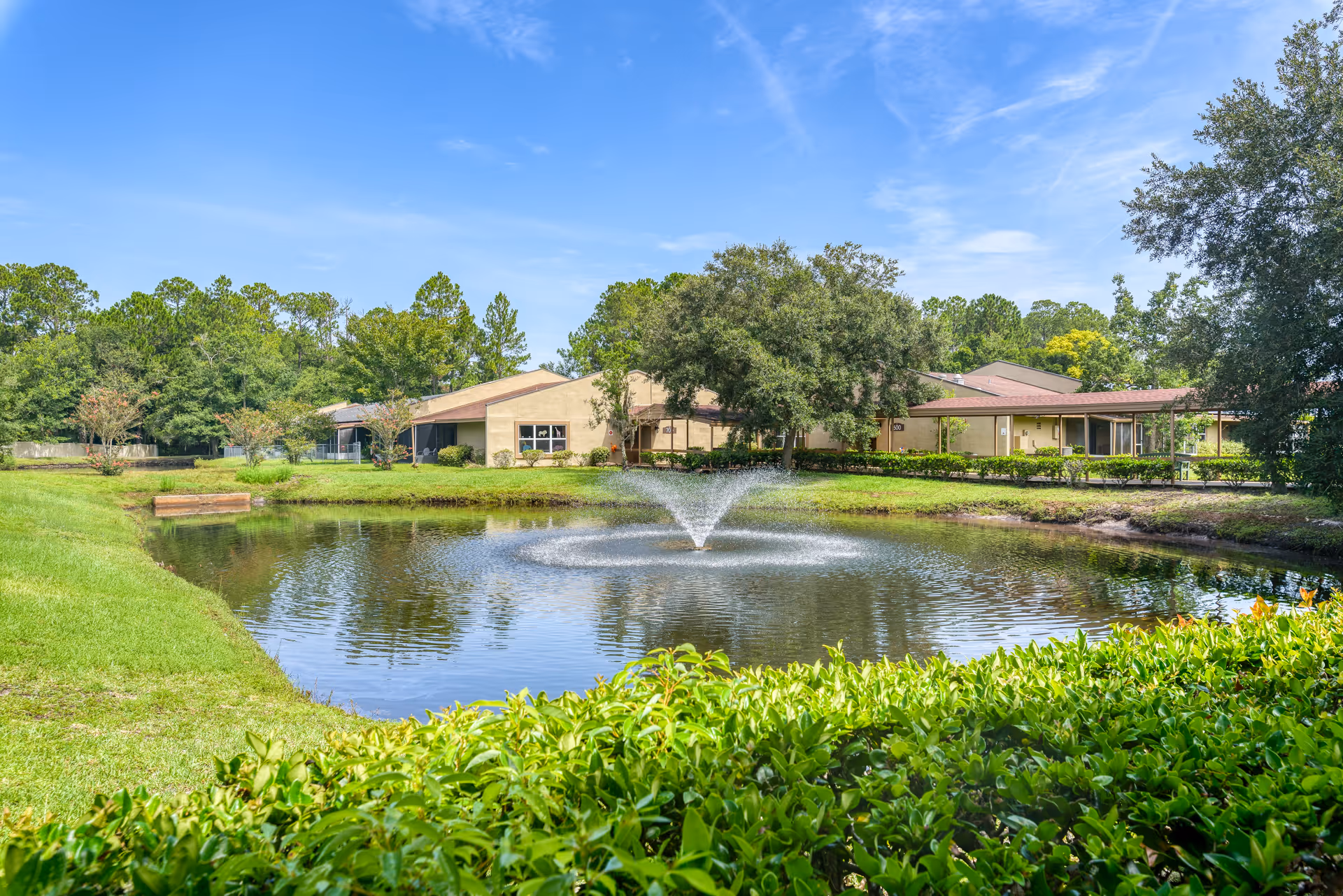 Landscaped pond with a central fountain in front of low-rise buildings and trees under a blue sky.