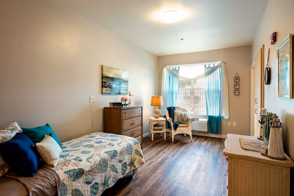 A cozy bedroom in a senior living facility featuring a single bed with patterned bedding and multiple pillows, a wooden dresser with decorative items, a wicker chair with a cushion and throw blanket near a window with blue curtains, a small side table with a lamp, and a wicker storage bench. The room has wood flooring and soft lighting.