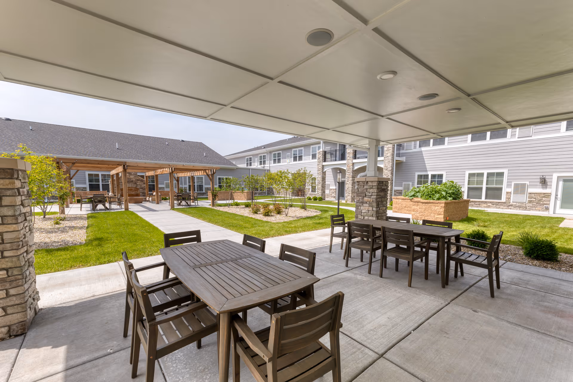 Outdoor covered patio area with wooden tables and chairs at Cedarhurst Senior Living of McHenry. The patio overlooks a landscaped courtyard with grass, small trees, and raised garden beds. In the background, there are pergolas and a two-story building with gray siding and stone accents.