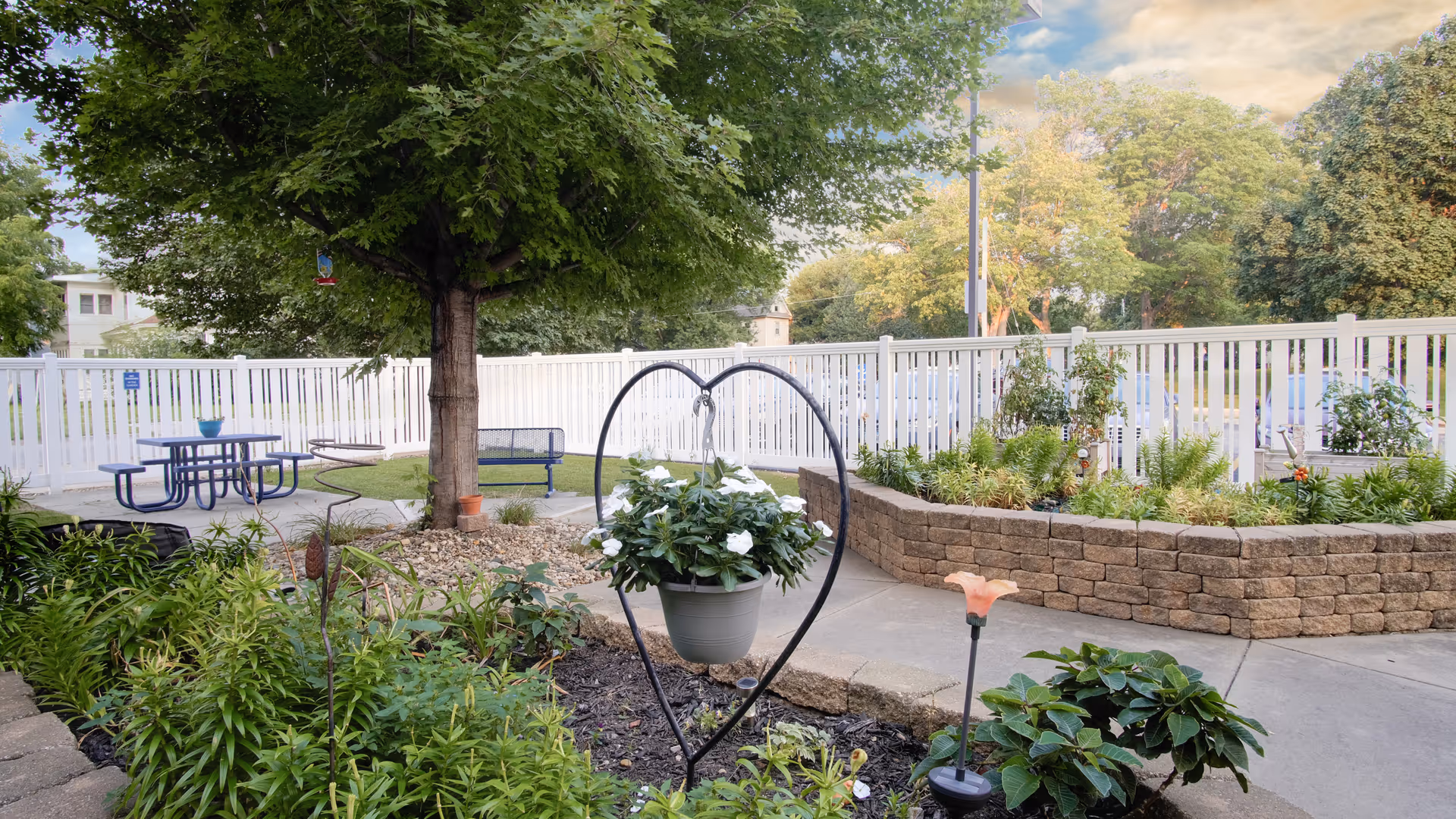 A courtyard garden with a heart-shaped metal hanger holding a potted plant, surrounding flowerbeds, a picnic table and benches next to a white fence.
