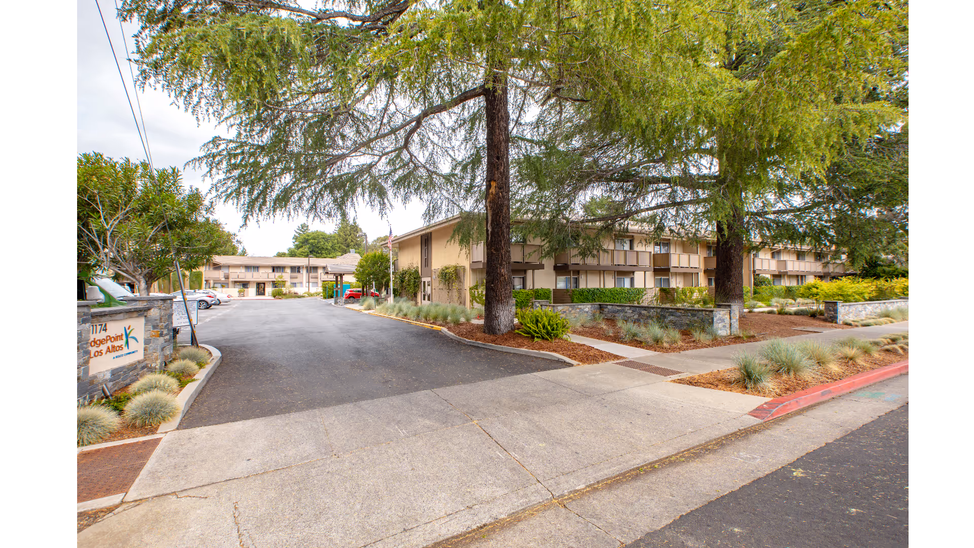 Entrance driveway to BridgePoint at Los Altos senior living facility with a stone sign on the left displaying the facility name and address. The driveway leads to a two-story building surrounded by trees and landscaped greenery under a cloudy sky.