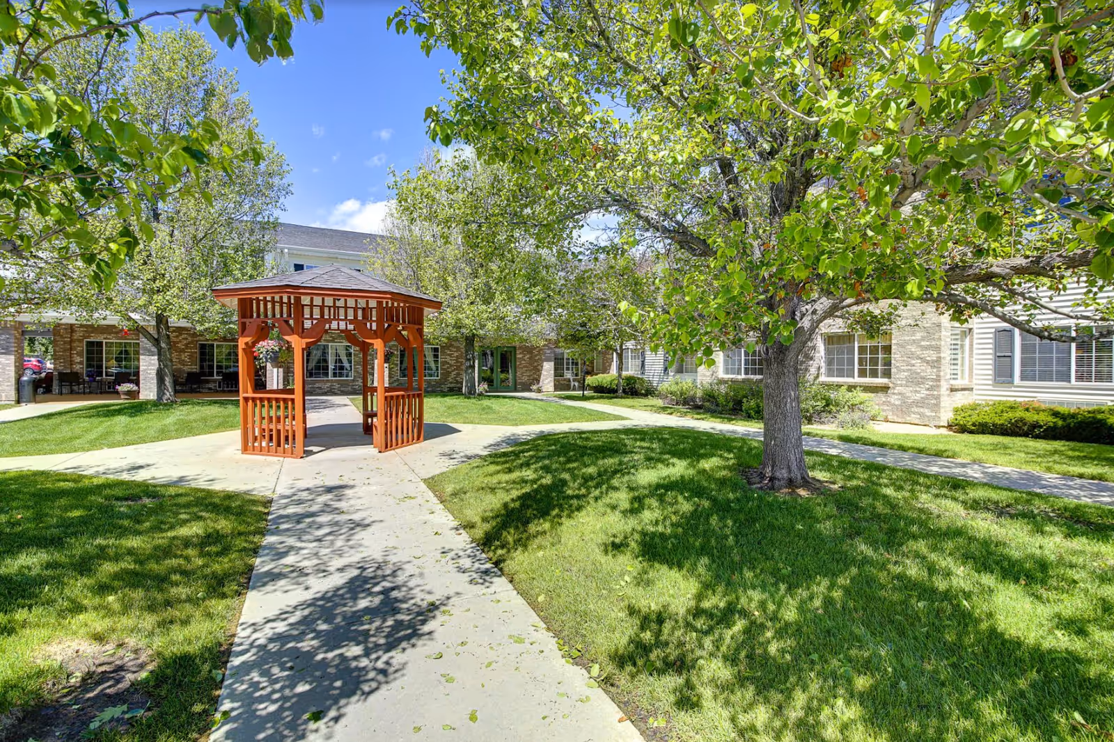 Outdoor courtyard area at Waneka Park Assisted Living featuring a red wooden gazebo in the center, surrounded by green grass, trees with green leaves, and a paved walkway leading to the gazebo. The building with large windows and a brick facade is visible in the background under a blue sky with some clouds.