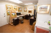 A small art studio room with several framed paintings on the walls and an easel holding a painting. The room has a wooden floor, a desk with a computer monitor, a chair, and a small sink with a red towel hanging nearby.