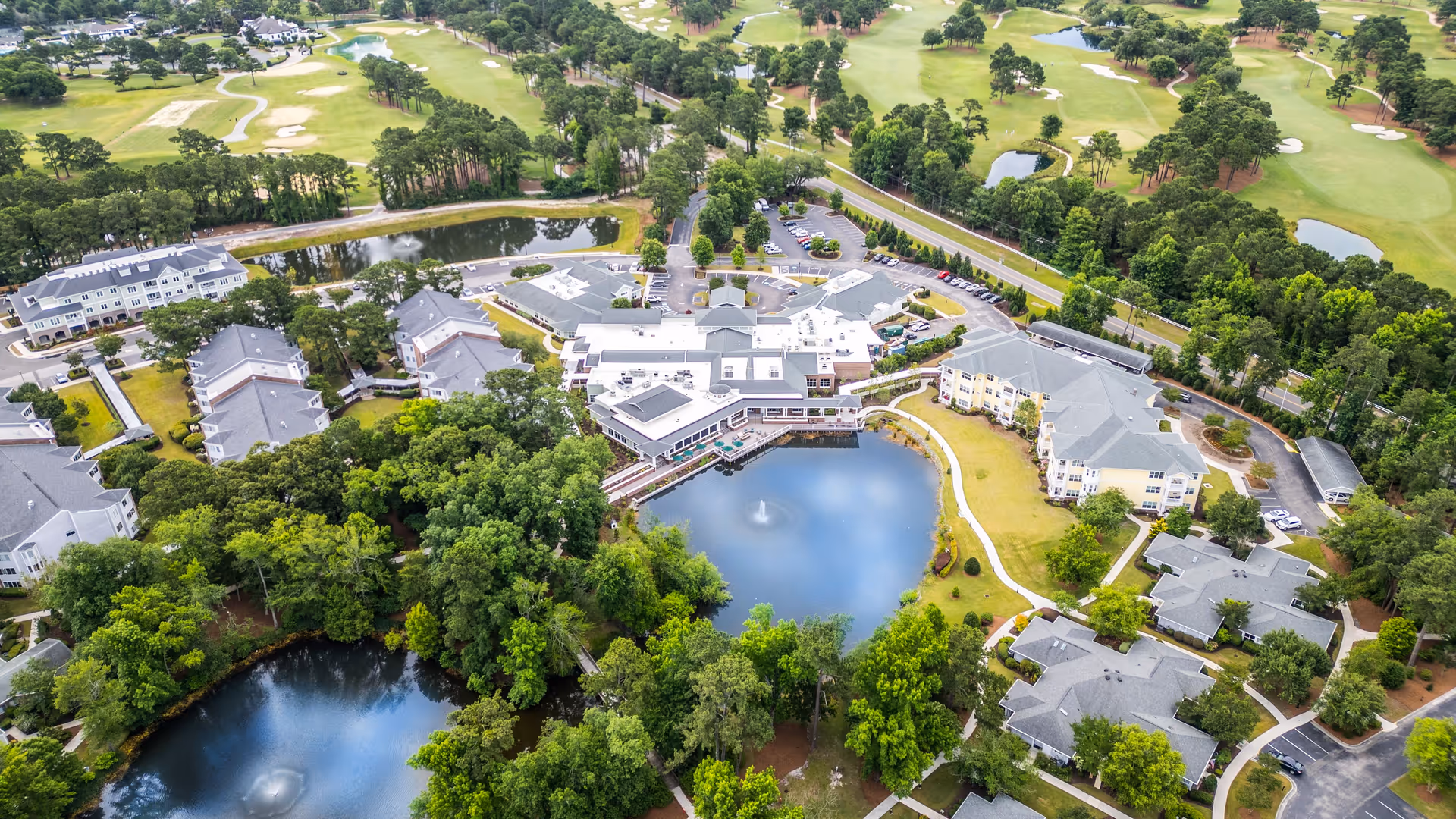 Aerial view of Porters Neck Village senior living facility showing multiple buildings surrounding a pond with a fountain, lush green trees, walking paths, parking areas, and a golf course in the background.