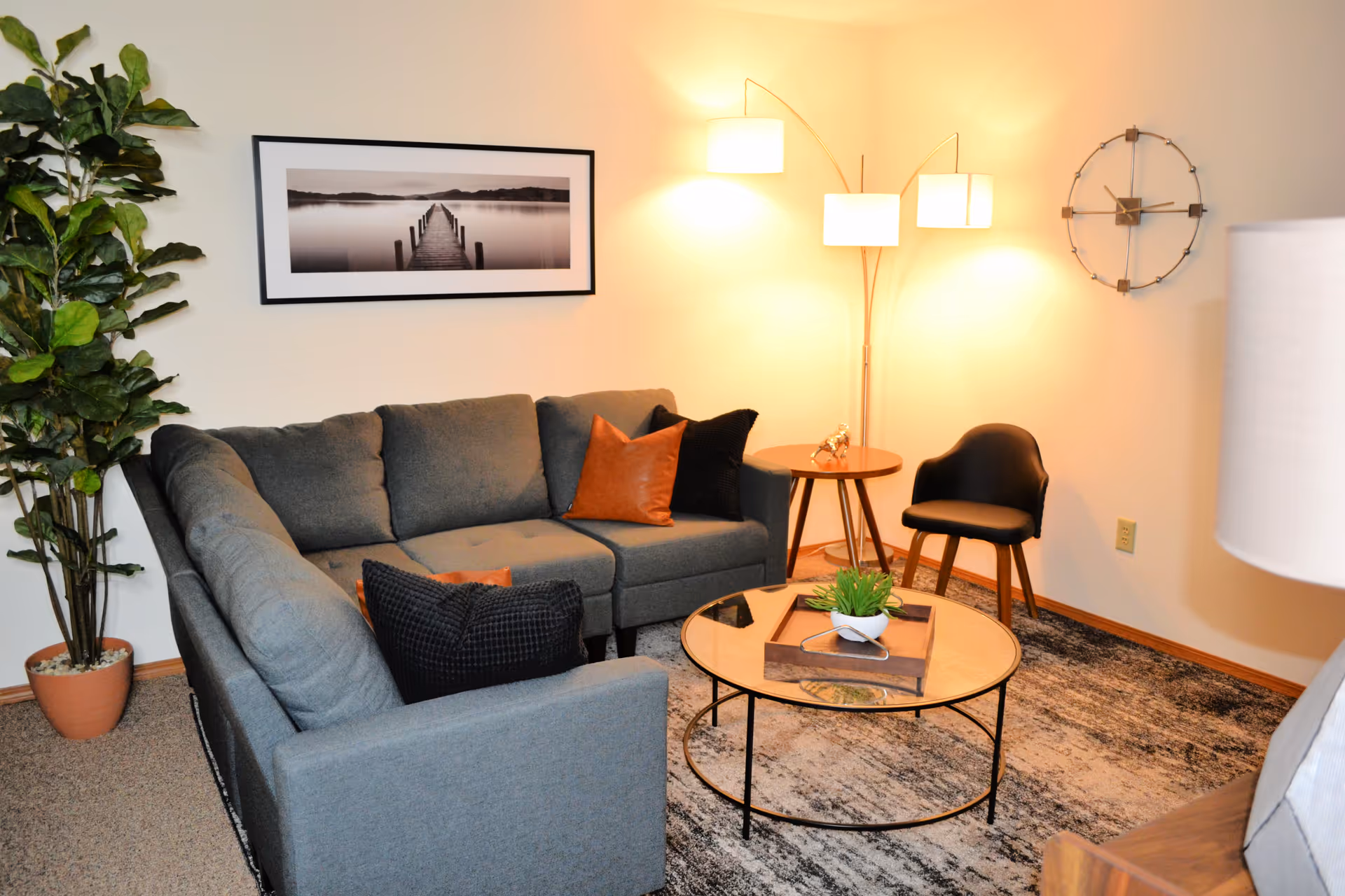 A cozy living room with a gray sectional sofa adorned with black and brown pillows, a round glass coffee table with a small plant and tray, a black chair, a wooden side table with a decorative item, a tall floor lamp with three lights, a large potted plant, a wall clock, and a framed black and white photo of a pier extending into a calm lake.