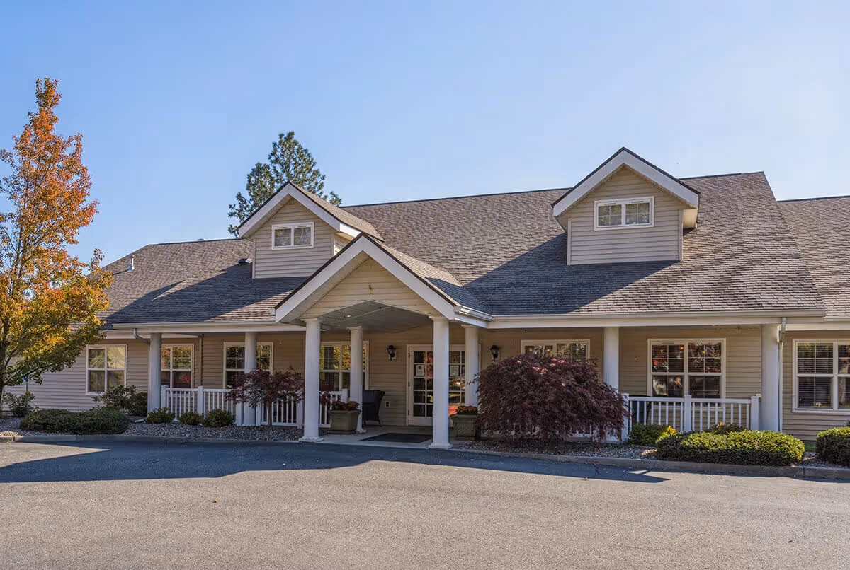 Front exterior of a single-story senior living building with a covered entrance supported by white columns, dormer windows, and landscaped shrubs under a clear blue sky.